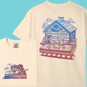 May include: Cream-colored t-shirt featuring a mushroom and train graphic on the front and a whimsical illustration on the back. The back design includes a building with the text "JURMO & TAPAL STATION" and frog characters playing music. The graphics are in blue and red.