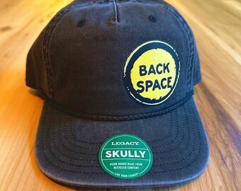 May include: A charcoal gray baseball cap featuring a circular yellow patch with the black text BACK SPACE. A black rope trim sits above the brim. A green circular sticker on the brim reads LEGACY SKULLY VISOR BOARD MADE FROM RECYCLED CONTENT.