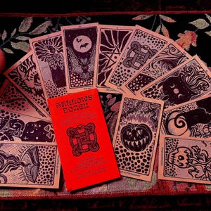 May include: A deck of tarot cards with black ink illustrations on tan paper, fanned out around a bright red box labeled Hallows Dozen. The cards feature imagery of bats, pumpkins, skulls, and cats. Two lit candles sit on either side of the cards.