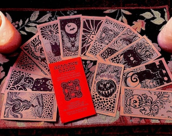 May include: A deck of tarot cards with black ink illustrations on tan paper, fanned out around a bright red box labeled Hallows Dozen. The cards feature imagery of bats, pumpkins, skulls, and cats. Two lit candles sit on either side of the cards.