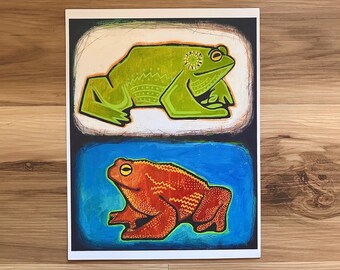 May include: A colorful art print featuring two stylized frogs. The top frog is green with yellow accents, set against a white background. The bottom frog is orange, set against a blue background. The artwork is framed in a white border.