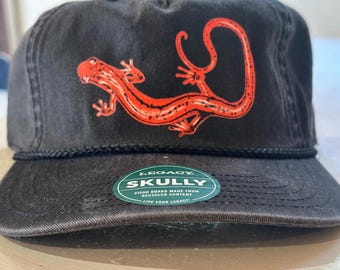May include: A black baseball cap featuring a bright orange salamander graphic on the front panel. The cap includes a black braided cord across the base of the crown and a green circular sticker on the brim that reads LEGACY SKULLY.