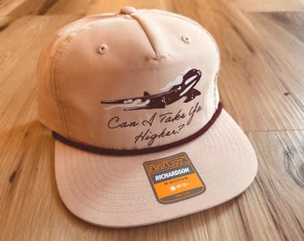 May include: A tan baseball cap with a flat brim, featuring a brown orca graphic and the text "Can I Take Ya Higher?" A maroon rope detail accents the brim. The cap includes a Richardson R-Active SPF 50+ tag.