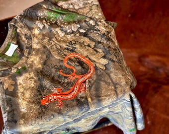 May include: A short-sleeve t-shirt featuring a camouflage pattern in shades of brown, tan, and green. A bright orange salamander with black spots and a dark stripe along its back is printed on the front. The text "REALTREE APX" is visible near the collar.