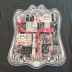 May include: A dark grey t-shirt featuring a white graphic print of an open alligator mouth. Inside the mouth is a detailed illustration of a modular synthesiser system with various black and white control panels and tangled red and yellow patch cables.