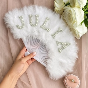 May include: A white feather fan with the name "JULIA" spelled out in pearl-studded letters. The fan is held open, revealing its pleated design. A ring box and white roses are in the background.