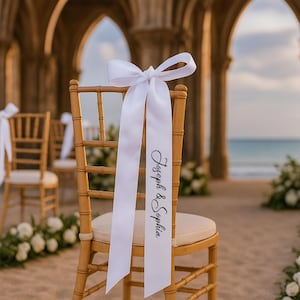 Op de afbeelding: Een houten stoel met een crèmekleurig kussen is versierd met een wit satijnen lint dat in een strik is geknoopt. Op het lint staan de namen "Joseph & Sophia" in sierlijke letters, wat duidt op een bruiloft of speciale gelegenheid.