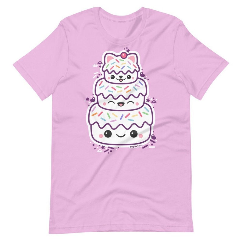 May include: A light pink t-shirt featuring a stacked cartoon cake with a cat-like face on top. The cake has white frosting, colorful sprinkles, and smiling faces. The text "sugarhai" is at the bottom.