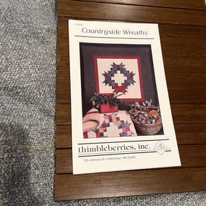May include: A printed paper with the title "Countryside Wreaths" and the text "thimbleberries, inc." is displayed on a brown wooden surface. The paper features images of quilts and wreaths. The background is a textured, blue-gray fabric.
