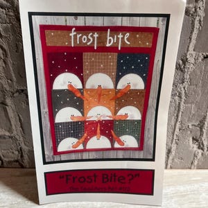 May include: A handmade quilt pattern titled "Frost Bite?" features a patchwork design with a red border and a variety of fabric squares. The quilt design includes white semicircles with orange carrot-like shapes. The text "frost bite" is written in white script.