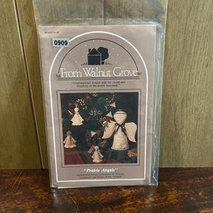 May include: A clear plastic package containing a craft pattern booklet titled From Walnut Grove Prairie Angels. The cover features a graphic of quilted angel ornaments and a garland, with text describing the contents as quilted and pieced items.