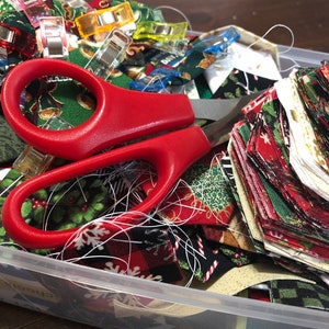 May include: A clear plastic storage container filled with colorful fabric scraps, red scissors, and a variety of colorful clothespins.