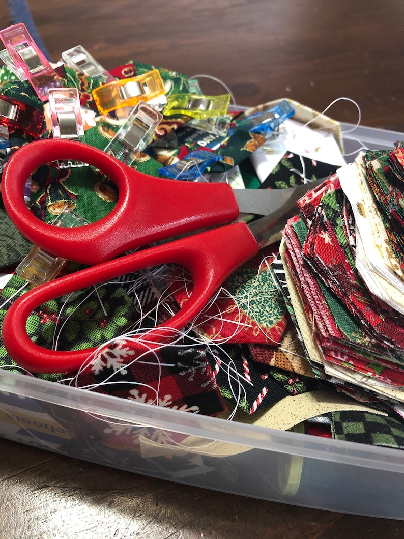 May include: A clear plastic storage container filled with colorful fabric scraps, red scissors, and a variety of colorful clothespins.