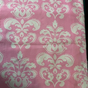 May include: A fabric featuring a repeating damask pattern in off-white against a solid light pink background. The design consists of symmetrical, ornate floral and leaf-like motifs arranged in a vertical grid, creating a continuous textile print.