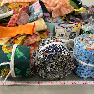 May include: A collection of colorful fabric scraps, some rolled and tied with white string. The fabrics feature floral, geometric, and abstract patterns in various colors. A measuring tape is visible at the bottom.