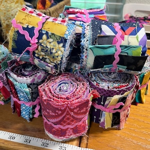May include: A collection of colorful fabric bundles, some rolled and others stacked, tied with pink ribbon. The fabrics feature floral, geometric, and abstract patterns in various colors, including blue, yellow, and red. A measuring tape is visible.