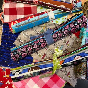May include: A collection of colorful fabric scraps, some rolled up and tied with ribbon, others laid flat. The fabrics feature various patterns, including gingham, floral, and geometric designs.