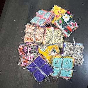 May include: A stack of colorful fabric squares, each tied with a ribbon, are arranged in a pile on a dark gray surface. The fabric squares are made of various patterns and colors, including floral, geometric, and abstract designs.