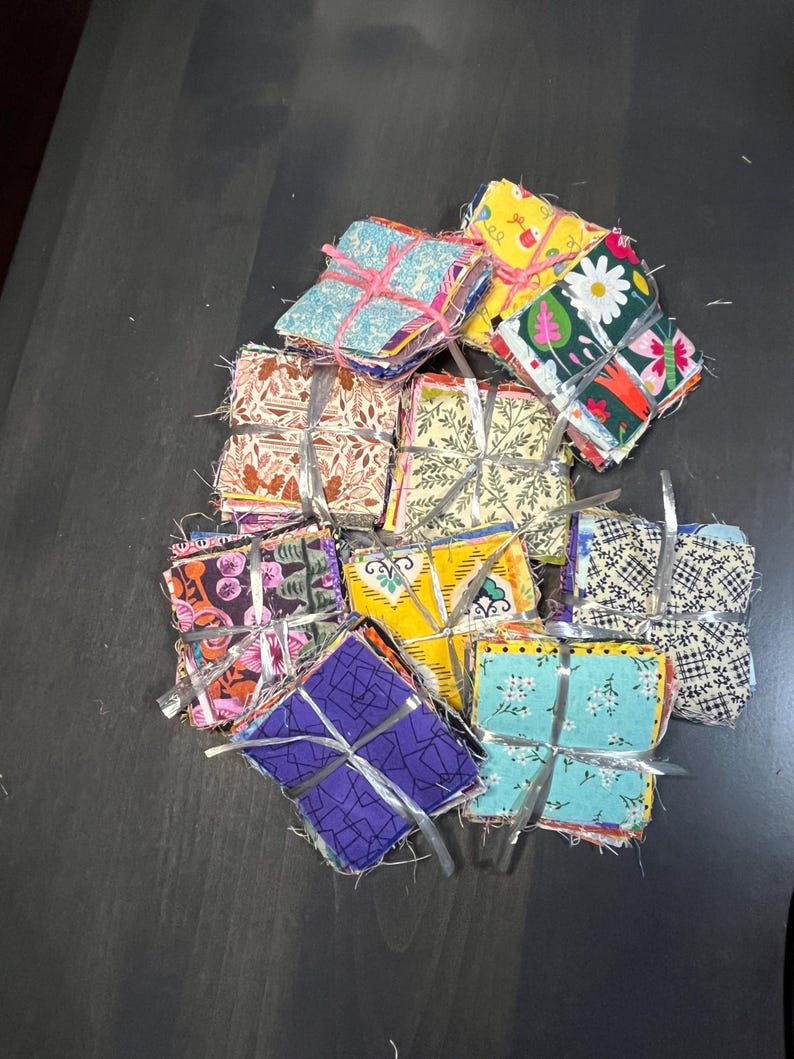 May include: A stack of colorful fabric squares, each tied with a ribbon, are arranged in a pile on a dark gray surface. The fabric squares are made of various patterns and colors, including floral, geometric, and abstract designs.