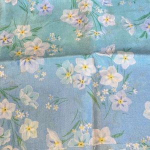 May include: Close-up of a light blue fabric with a floral pattern. The design features white flowers with yellow centers and light blue petals, along with smaller white and yellow flowers and green stems.