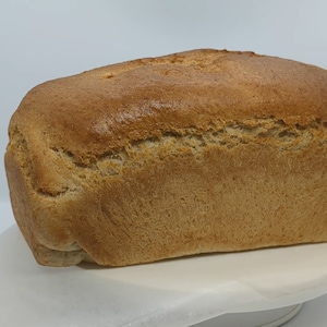 May include: A loaf of freshly baked bread, golden brown in color, with a slightly rough texture. The bread is sitting on a white surface.