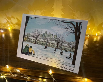 Może przedstawiać: Aquarela przedstawiająca śnieżną scenę miejską z ludźmi spacerującymi po śniegu. Ilustracja znajduje się na białej kartce z napisem "The Christmas Walk" napisanym czarnym kolorem na dole.