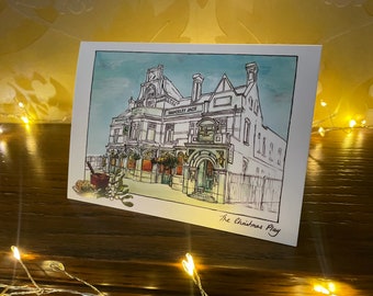 Może przedstawiać: Ręcznie rysowana ilustracja budynku z napisem "Drunken Jack" nad wejściem. Budynek jest narysowany w czerni i bieli na tle jasnoniebieskiego nieba. Ilustracja znajduje się na białej kartce z napisem "The Christmas Play" na dole.