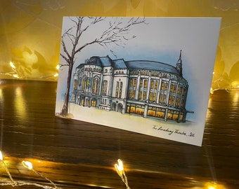 Może przedstawiać: Aquarela przedstawiająca Broadway Theatre w Saratoga Springs, Nowy Jork. Budynek jest przedstawiony w jasnoniebieskiej i białej kolorystyce z zaokrąglonym dachem i wysoką wieżą. Ilustracja jest umieszczona na tle jasnoniebieskiego nieba z kilkoma drzewami w tle.