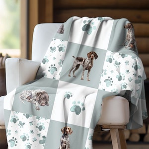 May include: A soft, white and light blue throw blanket with a repeating pattern of paw prints and German Shorthaired Pointer dogs. The blanket is draped over a white armchair.