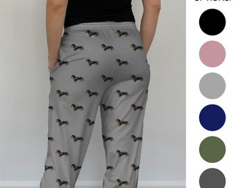May include: Gray pajama pants with a repeating pattern of dachshund dogs. The pants have a drawstring waist and pockets. The image also shows six color options: black, pink, gray, navy, olive, and dark gray. The text "6 COLOR OPTIONS!" is visible.