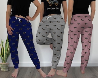Op de afbeelding: Drie pyjamabroeken in marineblauw, grijs en roze, elk bedrukt met illustraties van teckels. De afbeelding toont ook een zwart t-shirt met een teckelafbeelding en de tekst "Your Custom Text". De tekst "6 COLORS AVAILABLE!" en "ADD A MATCHING SHIRT!" zijn ook zichtbaar.