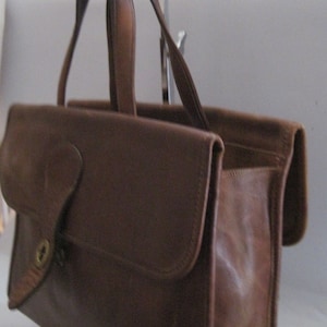 May include: A brown leather satchel handbag with a flap closure and a single handle. The bag has a brass lock and keyhole on the front.