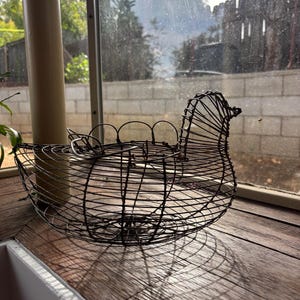 Op de afbeelding: Een draadmand in de vorm van een kip, gemaakt van donker metaal. De mand heeft een afgeronde basis en een gedetailleerd ontwerp van kop en staart. Het staat op een houten oppervlak bij een raam.