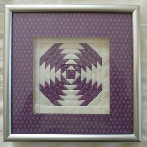 Puede incluir: Un bloque de colcha morado y blanco enmarcado con un patrón geométrico. El bloque de colcha está enmarcado en un marco plateado con un passe-partout morado estampado.