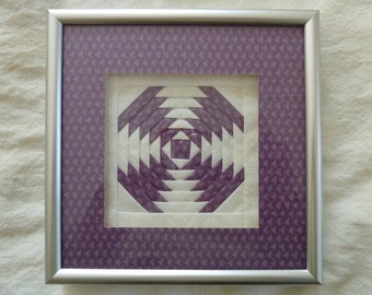 May include: A framed purple and white quilt block with a geometric pattern. The quilt block is framed in a silver frame with a purple patterned mat.