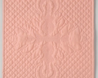 May include: A pink quilted fabric square with a raised, embroidered design in the center. The design is a cross shape with a floral pattern.