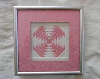 May include: A framed pink and white quilt block with a geometric pattern. The frame is silver and has a pink patterned mat.