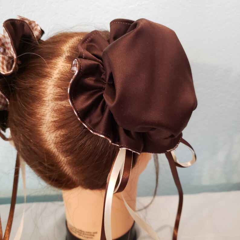 May include: A brown satin hair bun cover with white and brown ribbons. The bun cover is worn on a brown haired mannequin.