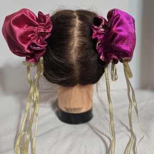 May include: Two hair accessories featuring deep magenta satin fabric gathered into rounded shapes, each accented with long, thin gold ribbons. The accessories are positioned on either side of a brown mannequin head with parted hair.