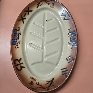 Op de afbeelding: Ovale keramische serveerschaal met een crèmekleurig midden en een bruine rand. De rand heeft gestileerde ontwerpen en symbolen in zwart en blauw. Het midden heeft een verhoogd bladerontwerp.