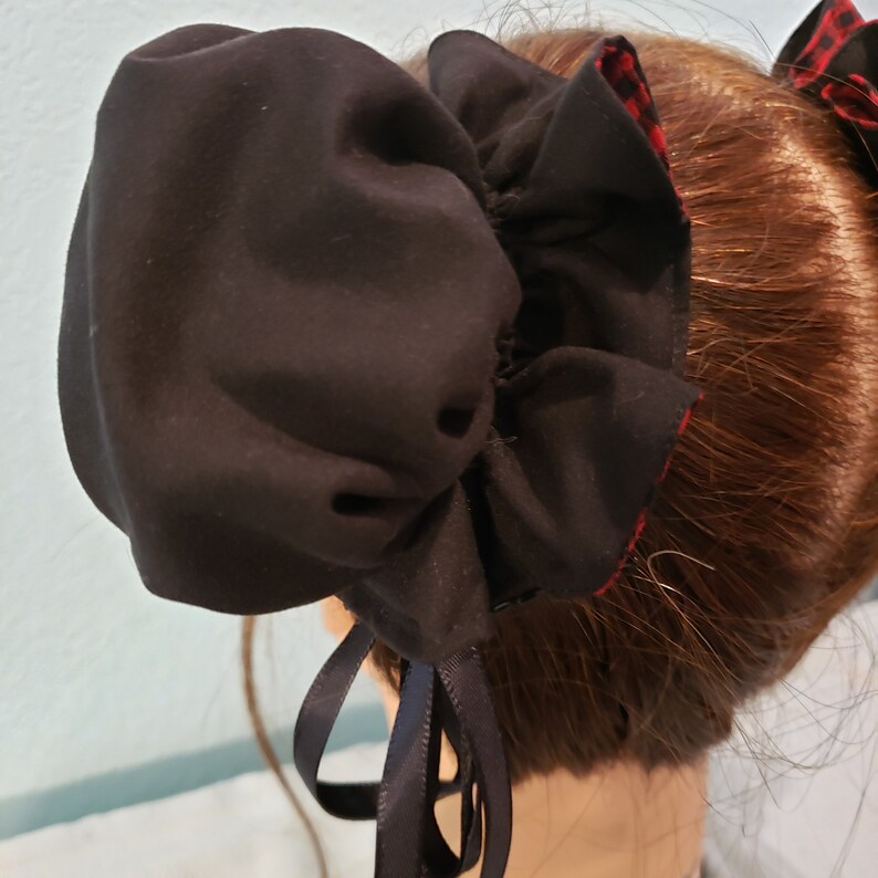 May include: Black fabric hair scrunchie with a ruffled edge and a black ribbon tie. The scrunchie is worn in a bun on a person's head.