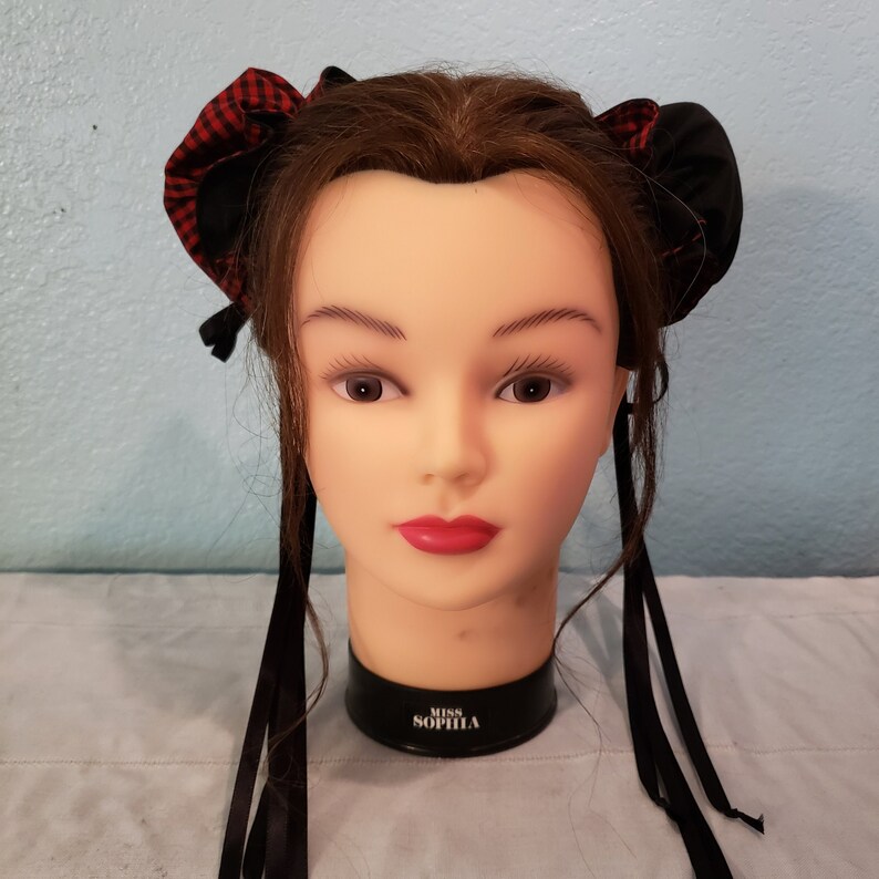 May include: A mannequin head with brown hair wearing a black and red plaid hair bow with long black ribbons.