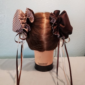 May include: Two brown and white gingham hair bows with long brown and white ribbons. The bows are tied on a brown wig head.