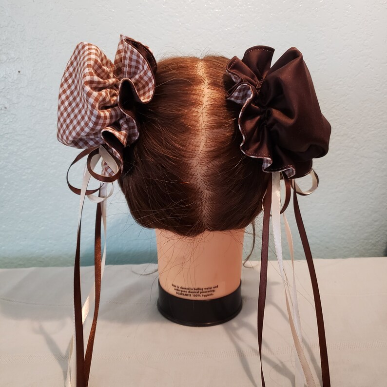 May include: Two brown and white gingham hair bows with long brown and white ribbons. The bows are tied on a brown wig head.