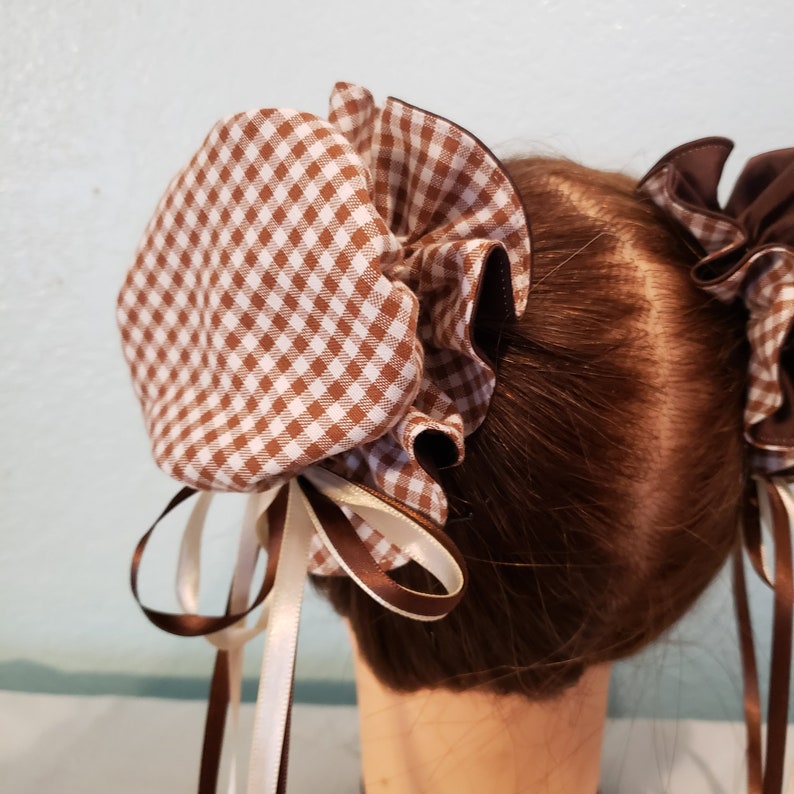 May include: Two brown and white gingham hair scrunchies with brown and white ribbon ties. The scrunchies are tied together in a bun on a brown wig.