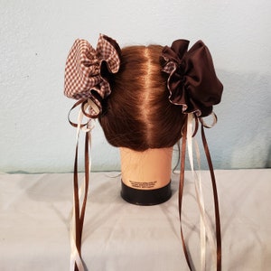 May include: Two brown and white hair bows with long brown and white ribbons. The bows are tied on a brown wig head.