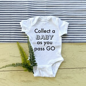 Può includere: Body da bambino bianco con la scritta "Collect a BABY as you pass GO" in nero. Il body è su una superficie giallo chiaro con un tessuto a righe blu e bianche sullo sfondo. Una felce verde è posizionata vicino al body.