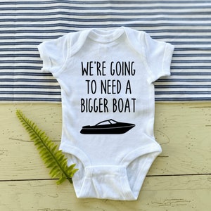 以下が含まれることがあります： 黒い文字で「We're going to need a bigger boat」と書かれた白いベビーロンパース。黒いスピードボートのシルエットが描かれている。