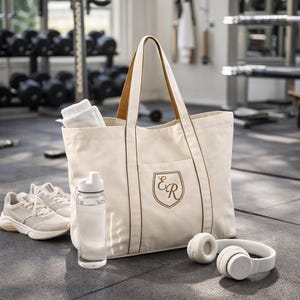 May include: Cream tote bag with brown handles and a monogrammed crest. White trainers, a water bottle, white headphones, and a white towel are also visible. The items are on a gym floor. The bag is suitable for carrying gym essentials.