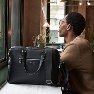May include: Black leather briefcase with silver zips and handles. The bag has a shoulder strap and the initials "Y.F" are embossed on the front. The briefcase is on a white surface.